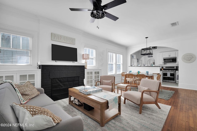 living room featuring a brick fireplace, light wood-type flooring, ceiling fan with notable chandelier, and ornamental molding