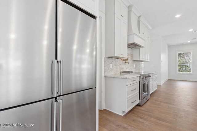 kitchen with custom exhaust hood, white cabinets, light wood-style floors, high quality appliances, and tasteful backsplash