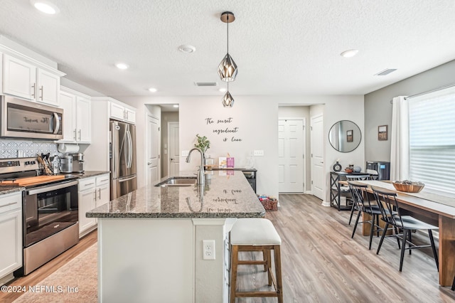 kitchen with stainless steel appliances, a center island with sink, sink, hanging light fixtures, and light wood-type flooring