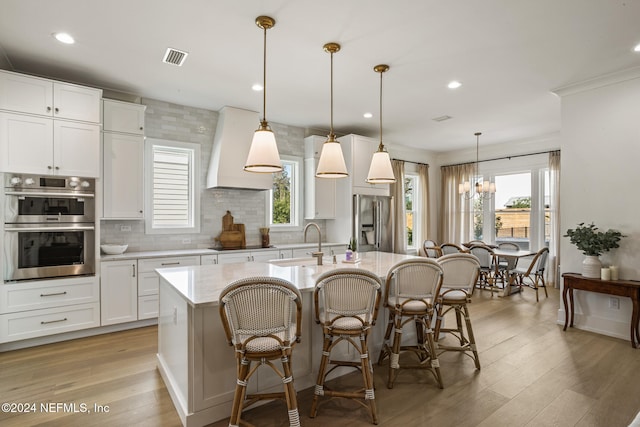 kitchen featuring a center island with sink, white cabinets, custom range hood, appliances with stainless steel finishes, and light wood-type flooring