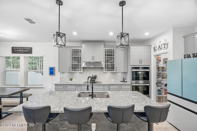 kitchen with an island with sink, stainless steel double oven, gray cabinets, backsplash, and pendant lighting