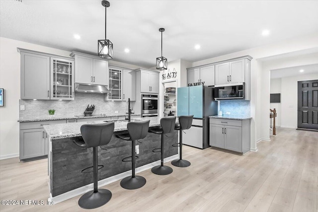 kitchen with a center island with sink, stainless steel appliances, gray cabinets, light stone countertops, and light hardwood / wood-style floors