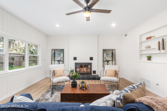 living room with built in shelves, ceiling fan, wooden walls, a tile fireplace, and light wood-type flooring