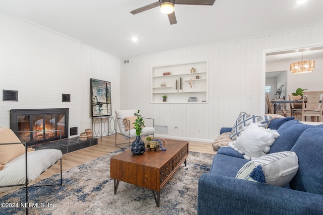 living room featuring built in shelves, wood walls, hardwood / wood-style flooring, ceiling fan, and a large fireplace