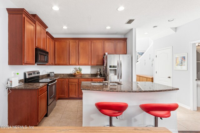 kitchen with stainless steel appliances, dark stone countertops, a textured ceiling, light tile patterned floors, and an island with sink