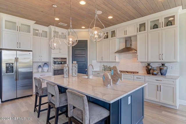 kitchen with an island with sink, appliances with stainless steel finishes, wall chimney exhaust hood, and hanging light fixtures