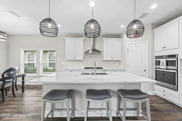 kitchen with wall chimney range hood, decorative light fixtures, a center island with sink, and white cabinets