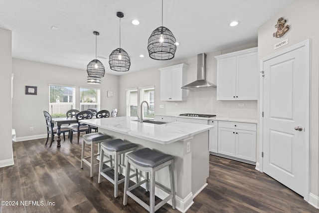 kitchen with a center island with sink, wall chimney exhaust hood, hanging light fixtures, gas stovetop, and white cabinets