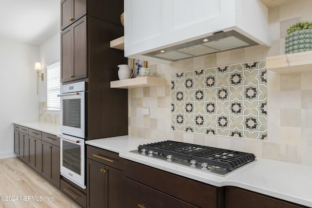 kitchen featuring stainless steel gas cooktop, backsplash, light hardwood / wood-style floors, dark brown cabinets, and custom exhaust hood