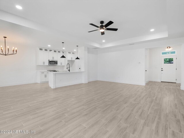 unfurnished living room featuring light wood-type flooring, a tray ceiling, sink, and ceiling fan with notable chandelier