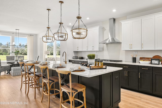 kitchen with a center island with sink, wall chimney exhaust hood, an inviting chandelier, decorative light fixtures, and white cabinets