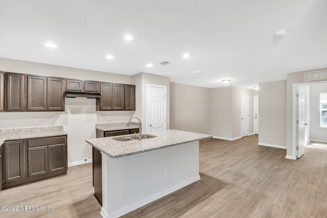 kitchen with sink, an island with sink, light stone countertops, dark brown cabinets, and light hardwood / wood-style flooring