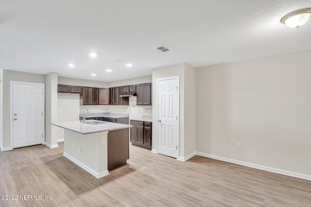 kitchen with light stone counters, a center island with sink, dark brown cabinetry, sink, and light hardwood / wood-style floors