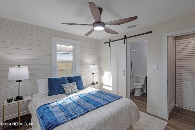 bedroom featuring ceiling fan, dark hardwood / wood-style floors, connected bathroom, a barn door, and wooden walls