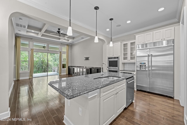 kitchen with dark stone counters, coffered ceiling, a center island with sink, appliances with stainless steel finishes, and white cabinetry