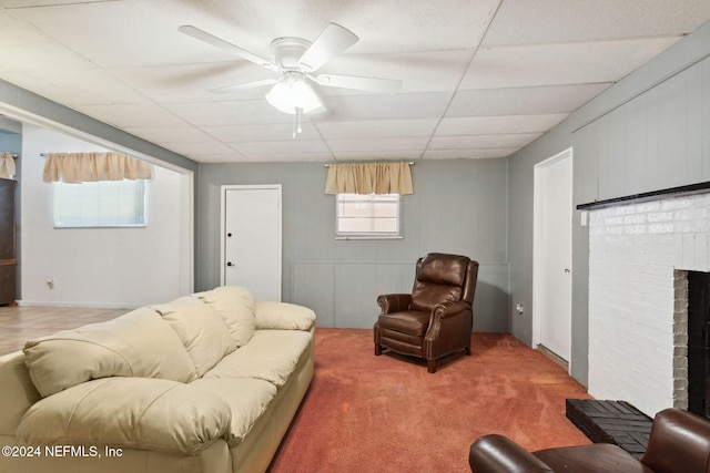 living room with a drop ceiling, carpet, a brick fireplace, and ceiling fan