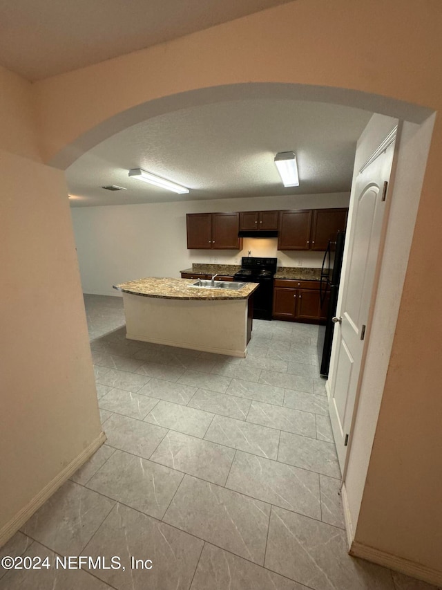 kitchen featuring black appliances, light tile patterned flooring, a center island with sink, dark brown cabinetry, and light stone countertops