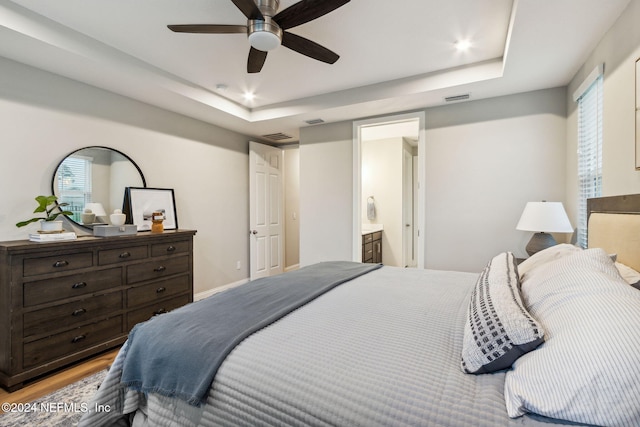 bedroom featuring ceiling fan, ensuite bath, a raised ceiling, and light wood-type flooring