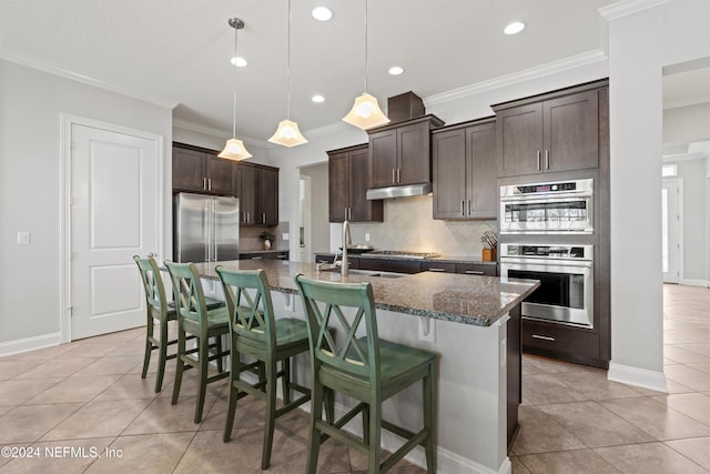 kitchen featuring appliances with stainless steel finishes, backsplash, dark brown cabinets, decorative light fixtures, and an island with sink