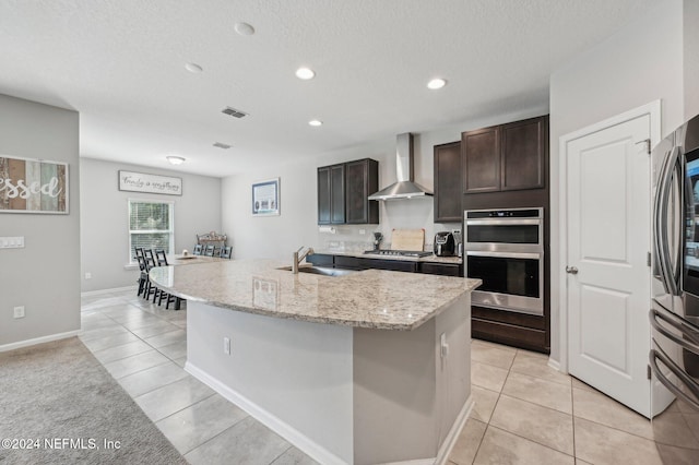 kitchen with stainless steel appliances, wall chimney range hood, light tile patterned floors, sink, and an island with sink