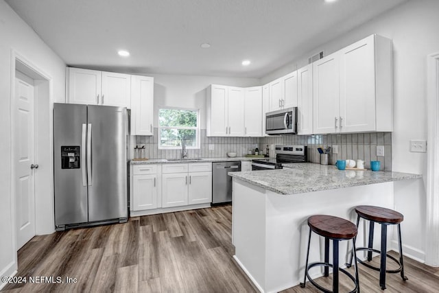 kitchen with kitchen peninsula, appliances with stainless steel finishes, a kitchen bar, hardwood / wood-style flooring, and white cabinetry