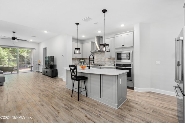 kitchen featuring hanging light fixtures, stainless steel appliances, wall chimney range hood, light hardwood / wood-style floors, and a kitchen island with sink