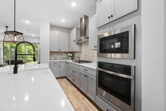 kitchen featuring gray cabinetry, wall chimney exhaust hood, hanging light fixtures, appliances with stainless steel finishes, and light wood-type flooring