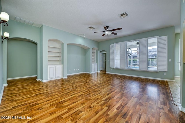 unfurnished living room with ceiling fan, built in shelves, a textured ceiling, and hardwood / wood-style floors