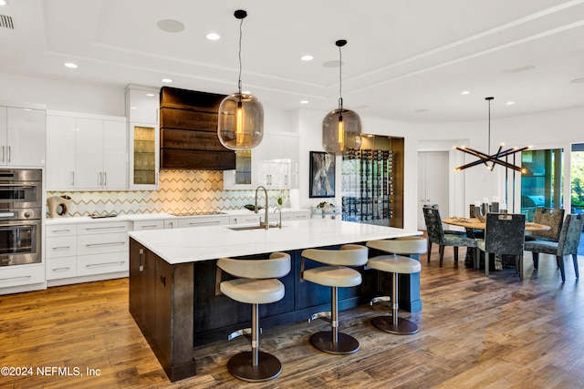 kitchen with sink, ventilation hood, double oven, hardwood / wood-style floors, and a kitchen island with sink