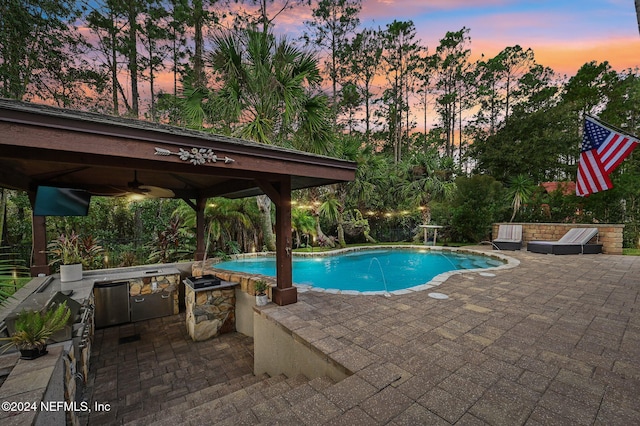 pool at dusk featuring an outdoor kitchen, a gazebo, ceiling fan, pool water feature, and a patio