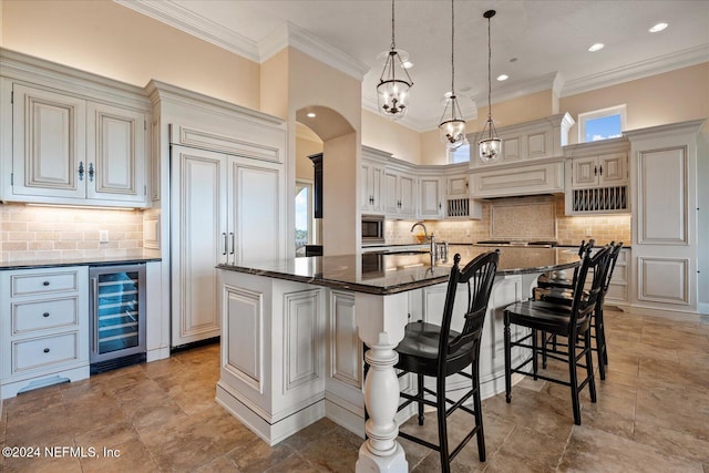 kitchen with dark stone counters, built in appliances, pendant lighting, an island with sink, and beverage cooler