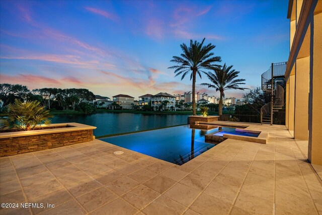 pool at dusk featuring a water view, a patio, and an in ground hot tub