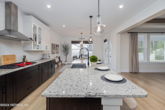 kitchen with a kitchen island with sink, wall chimney range hood, decorative light fixtures, light hardwood / wood-style flooring, and white cabinets