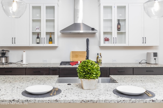 kitchen with light stone countertops, white cabinets, hanging light fixtures, and wall chimney range hood