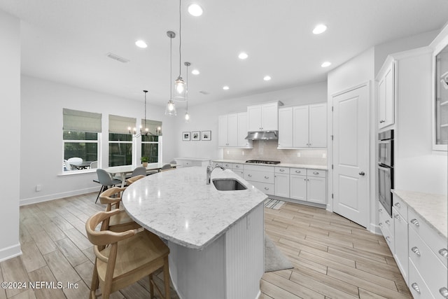 kitchen featuring light wood-type flooring, a kitchen island with sink, sink, white cabinetry, and hanging light fixtures