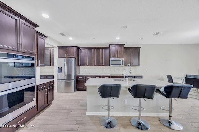 kitchen with appliances with stainless steel finishes, light wood-type flooring, backsplash, a breakfast bar, and an island with sink