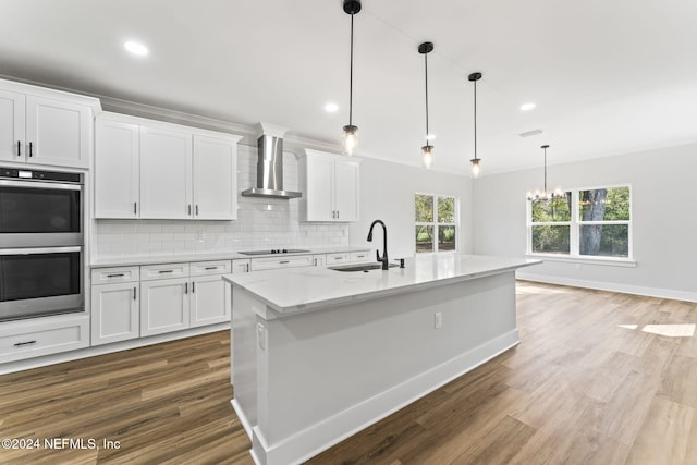 kitchen featuring hanging light fixtures, wall chimney range hood, double oven, a center island with sink, and white cabinets