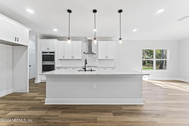 kitchen featuring an island with sink, stainless steel double oven, white cabinets, and decorative light fixtures