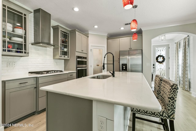 kitchen featuring gray cabinetry, wall chimney exhaust hood, stainless steel appliances, a kitchen island with sink, and sink