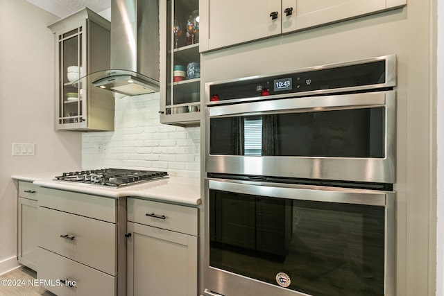 kitchen with gray cabinetry, wall chimney range hood, backsplash, and appliances with stainless steel finishes