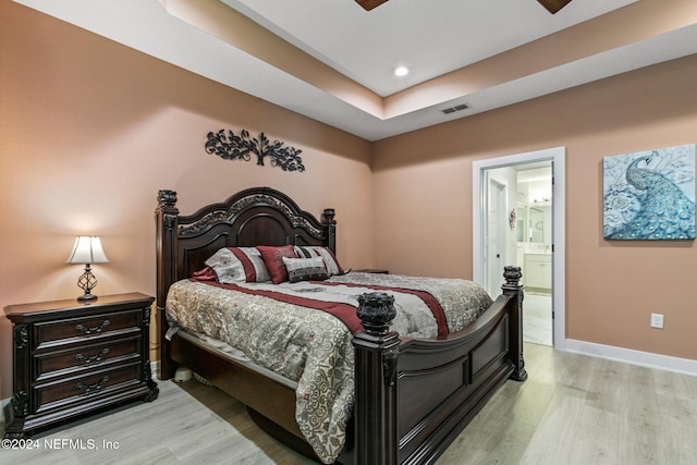 bedroom with light wood-type flooring, a tray ceiling, ensuite bath, and ceiling fan