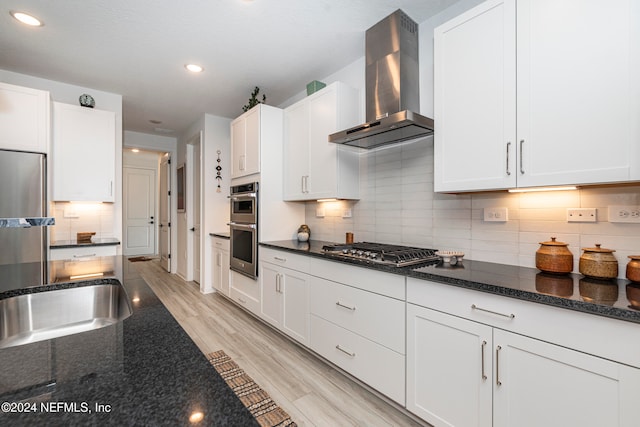 kitchen with tasteful backsplash, stainless steel appliances, wall chimney range hood, white cabinets, and light hardwood / wood-style floors