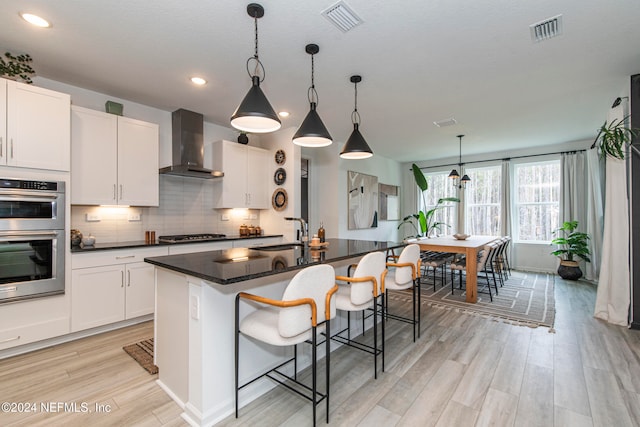 kitchen with white cabinets, an island with sink, decorative light fixtures, and wall chimney range hood