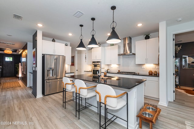 kitchen with pendant lighting, a center island with sink, wall chimney range hood, white cabinetry, and stainless steel appliances