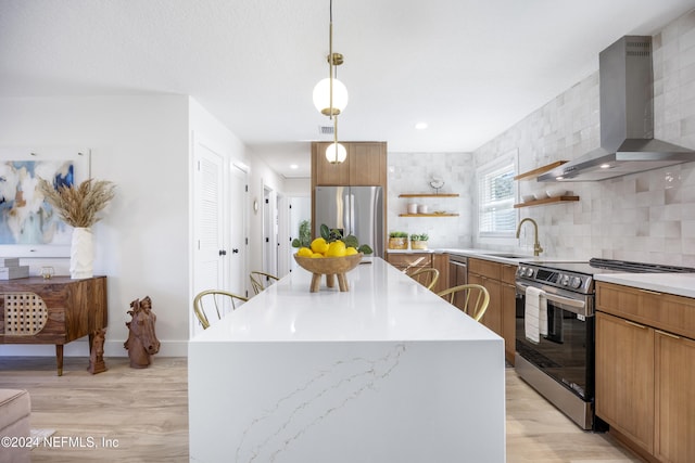 kitchen with pendant lighting, wall chimney range hood, light wood-type flooring, appliances with stainless steel finishes, and a kitchen island