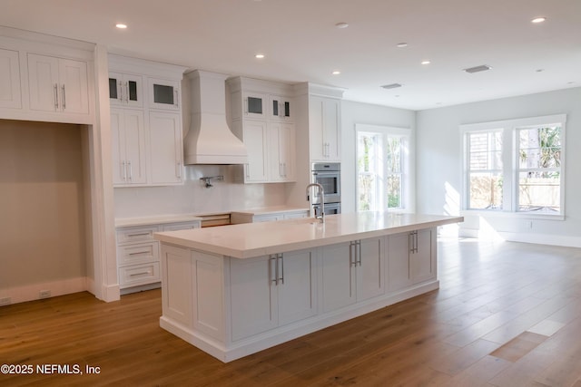 kitchen with custom range hood, a kitchen island with sink, glass insert cabinets, and light countertops