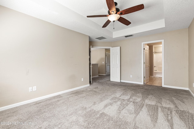 unfurnished bedroom featuring ensuite bath, a textured ceiling, light colored carpet, a raised ceiling, and ceiling fan