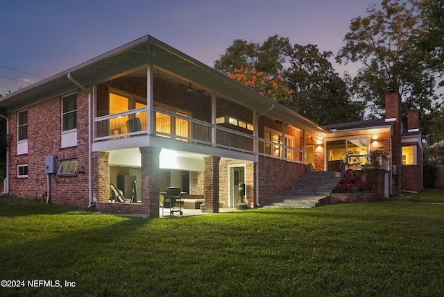 back house at dusk with a patio, a balcony, and a lawn