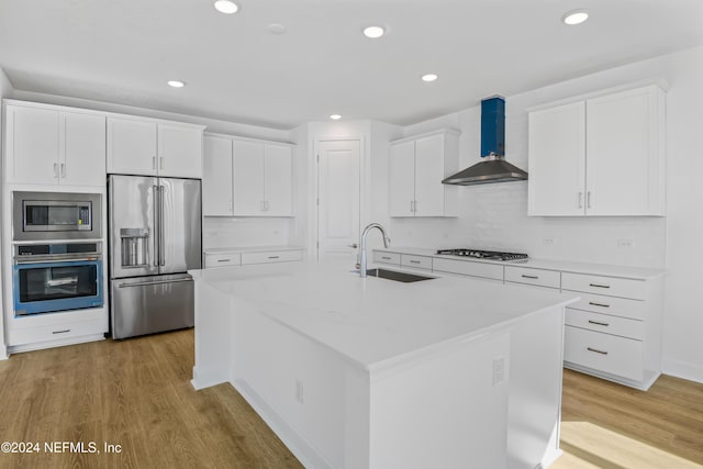 kitchen featuring appliances with stainless steel finishes, sink, wall chimney range hood, white cabinets, and an island with sink