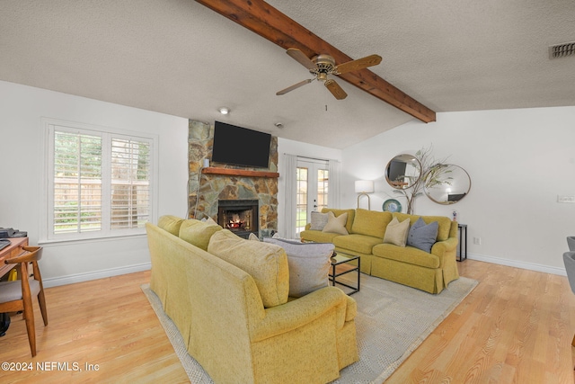 living room with lofted ceiling with beams, a stone fireplace, light hardwood / wood-style flooring, ceiling fan, and a textured ceiling